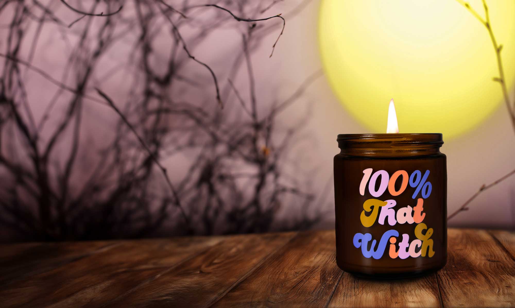 Photo of candle from Confia saying "100% that Witch" and with a composite background showing a full moon and bare branches with a warm glow.