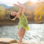 Dancer in passe pose on a rock with the sun streaming in behind on the water