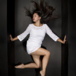 Dancer in white dress holding up on the walls as she jumps in an angled pose with her hair flying