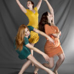 Dancers in yellow orange and teal dresses improvising poses on a grey background