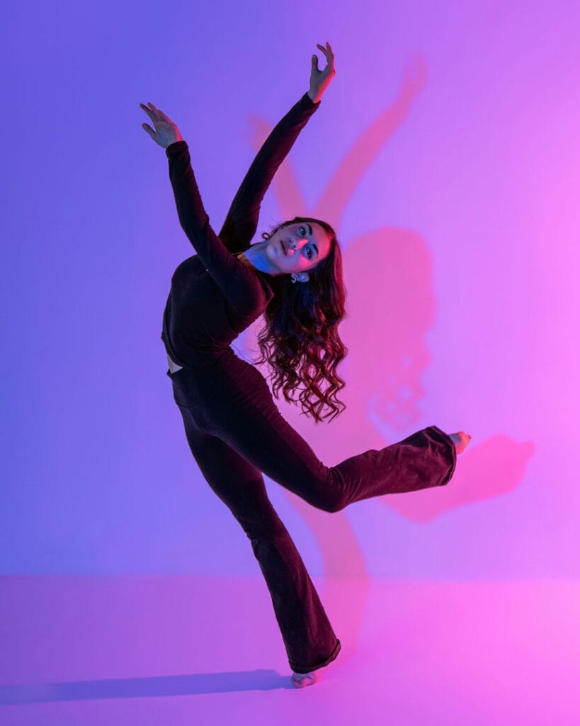 ballet dancer posed in attitude back leaning forward with arms raised on pink and purple