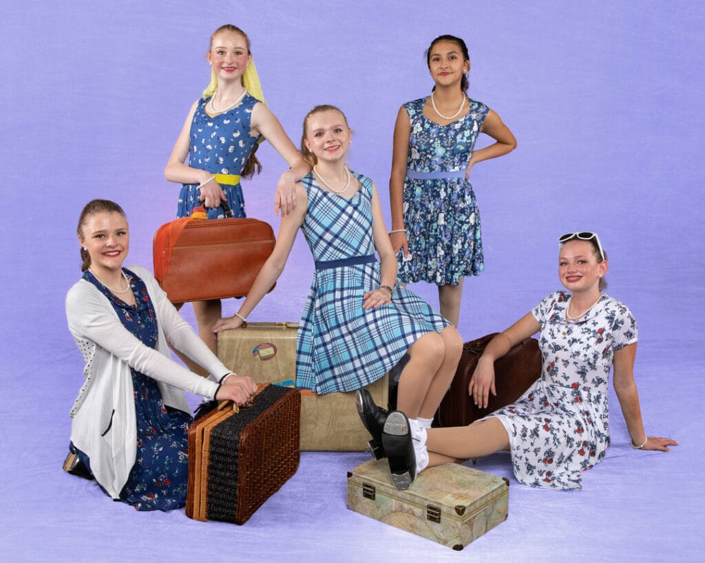 group of tap dancers posed in a staggered group with luggage on periwinkle