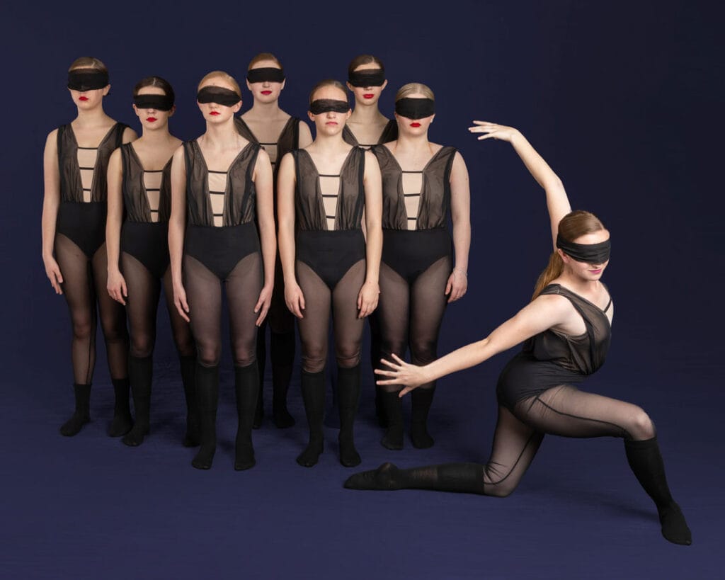 group of contemporary dancers posed in a simple tight group with one dancer breaking away in a lunge in front on blue