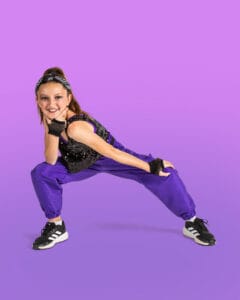 hip hop dancer posed in deep second lunge with one hand on knee and one under chin on magenta and purple background