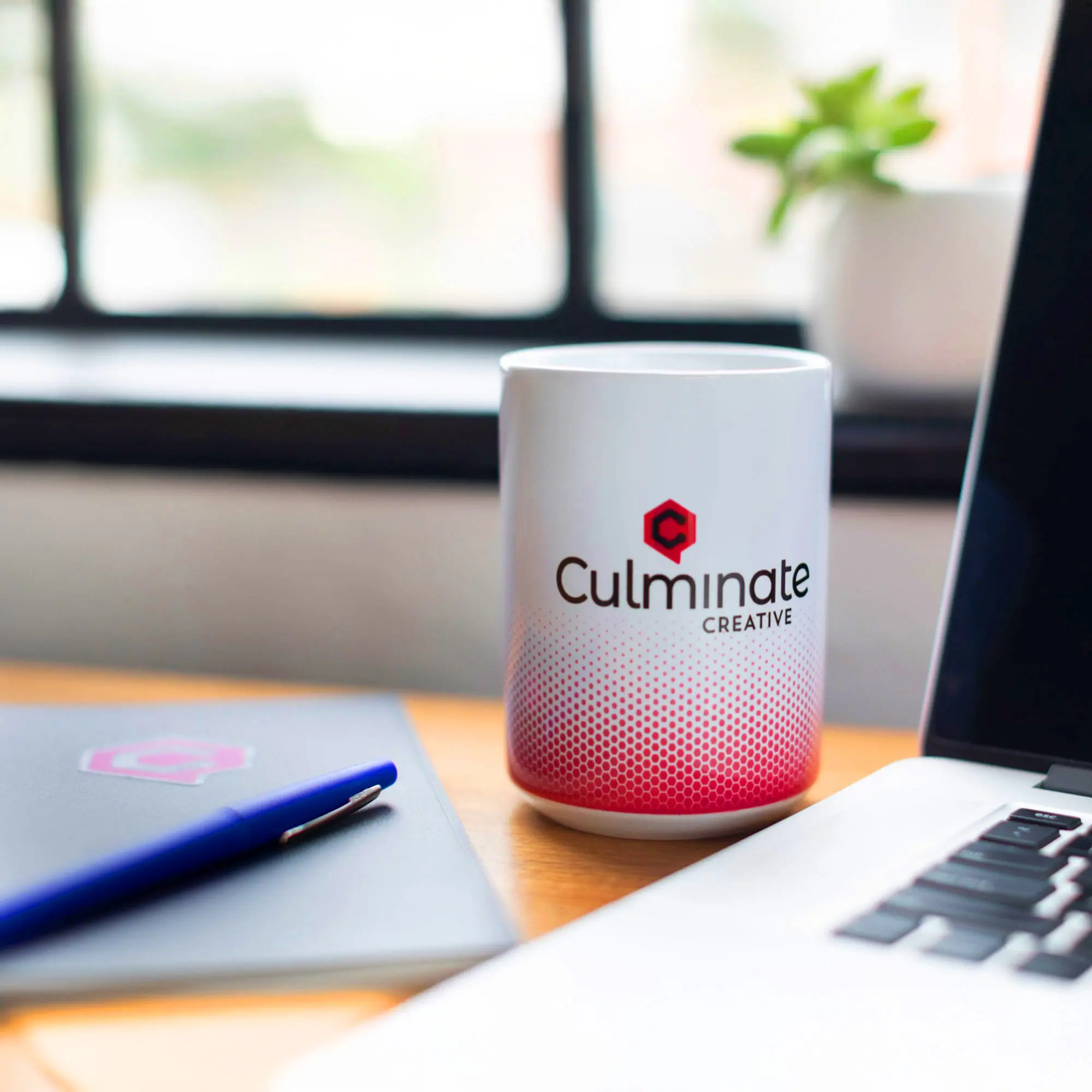 Laptop, notebook, pen, and branded mug on work desk with workspace background.