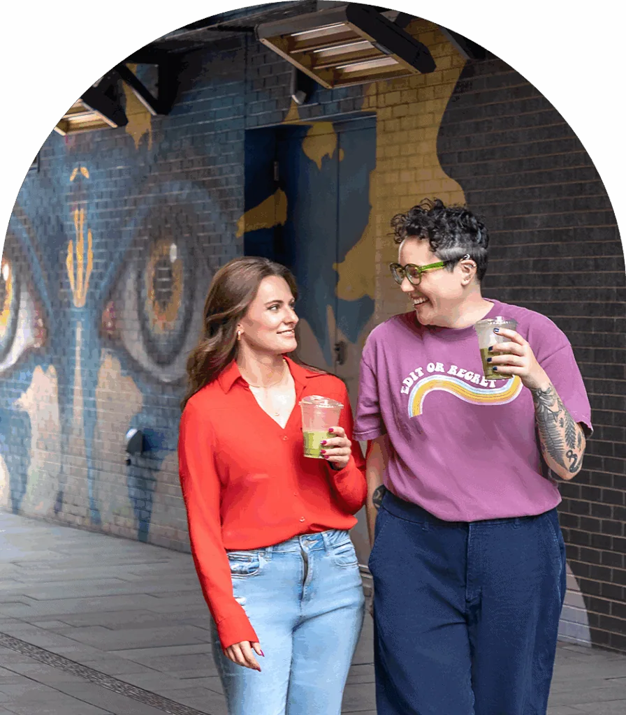 Vibrant mural art on brick wall with two women enjoying smoothies outdoors.