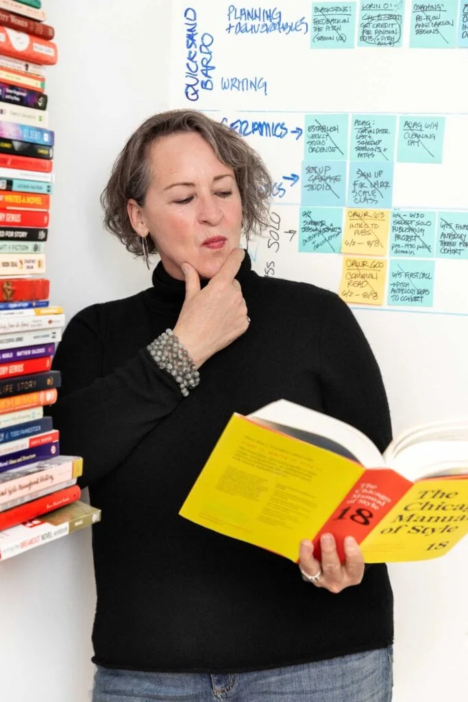 Creative woman reading a yellow and red book against a backdrop of bookshelves and planning whiteboard.